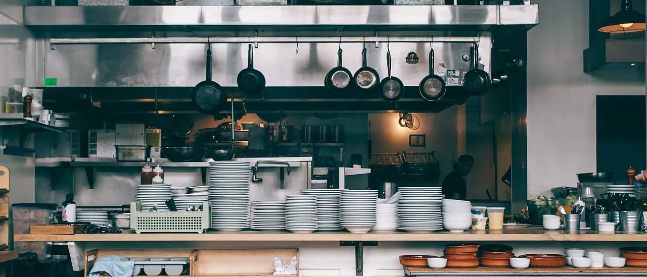 Stainless-steel commercial restaurant kitchen with preparation surfaces, stacked plates and cooking equipment.